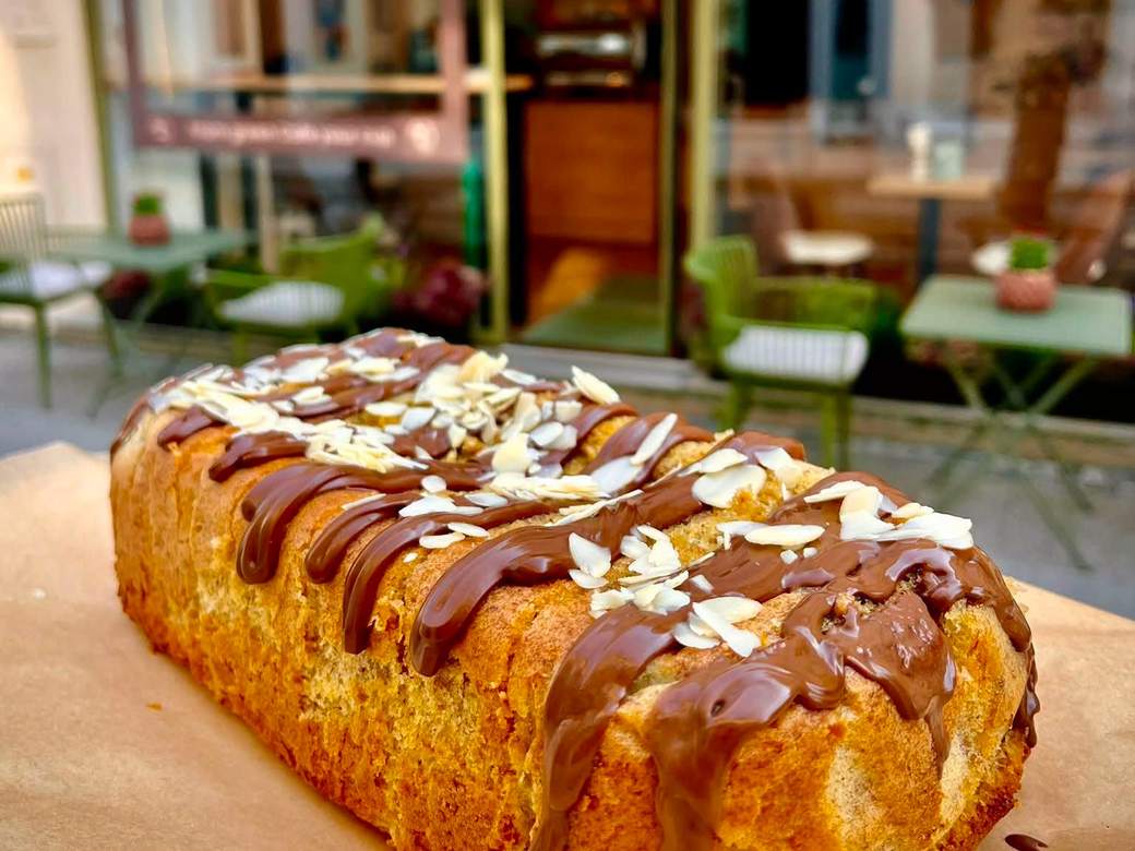 Frisch gebackener Bananenbrot-Laib mit Schokolade und Mandelblättchen, dahinter der Gastgarten vor dem Café