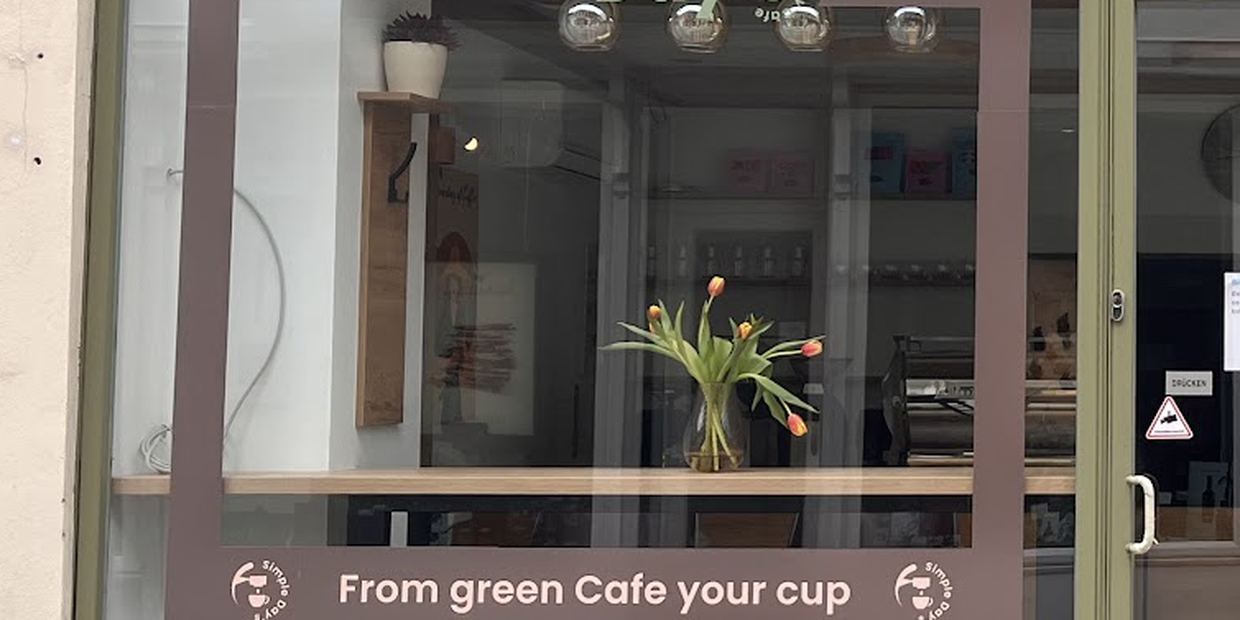Schaufensterscheibe von Simple Day&rsquo;s mit dem Schriftzug „From green Cafe your cup“ zwischen zwei Siegeln, dahinter Tulpen auf der Fensterbank