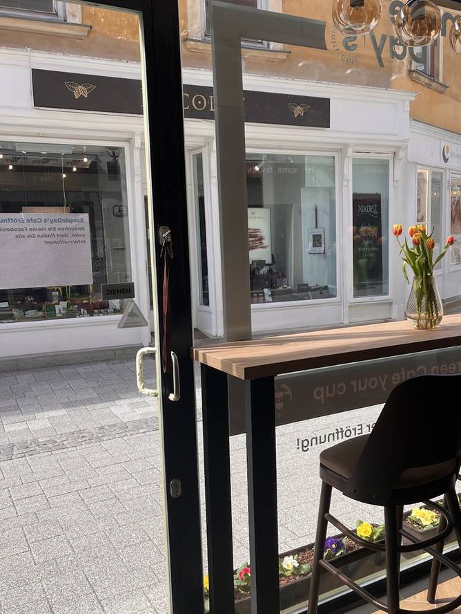 Blick aus dem Café durch die Glastür auf den Fensterbanktisch mit Tulpen, Barhocker und Blumenkisterl in der Theresiengasse
