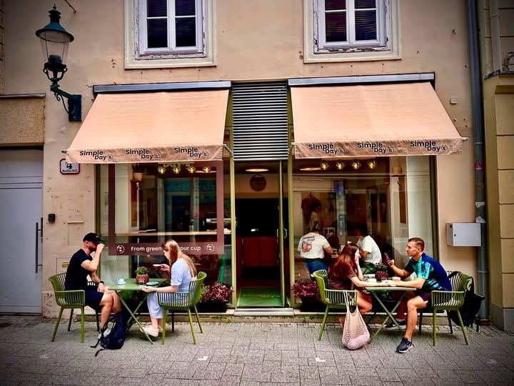 Gäste an den salbeigrünen Tischen im Gastgarten von Simple Day’s, über ihnen die beiden Markisen mit dem Schriftzug