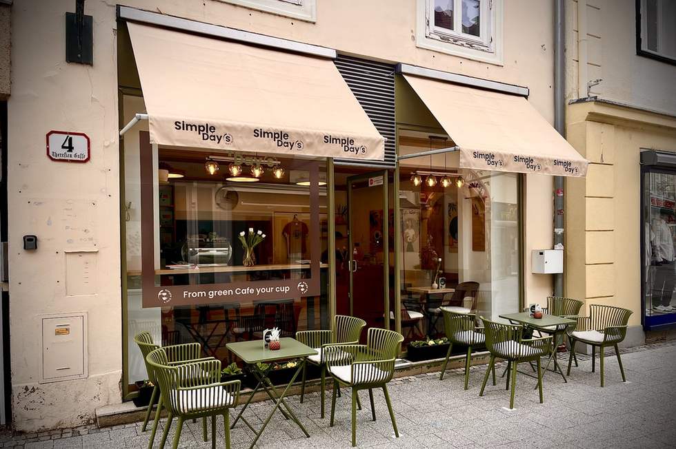 Die Fassade von Simple Day&rsquo;s Cafe in der Theresiengasse: helle Markise mit Schriftzug, salbeigrüne Bistrostühle und Tische in der Fußgängerzone