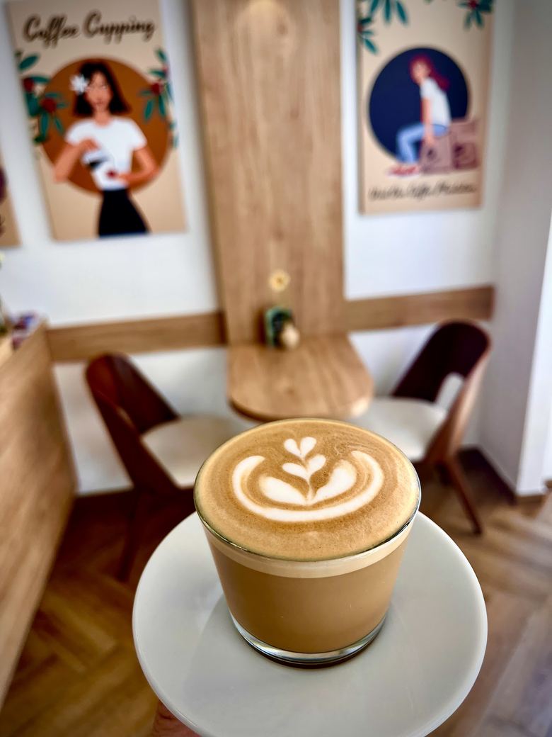 Flat White mit Rosetta-Latte-Art in einem Glas, dahinter die Illustrationen an der Wand des Cafés