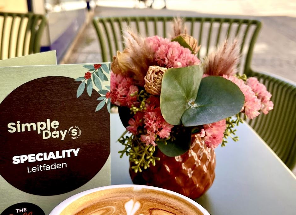 Der gedruckte Speciality-Leitfaden als Aufsteller auf einem Tisch im Gastgarten, daneben ein Trockenblumenstrauß und ein Cappuccino
