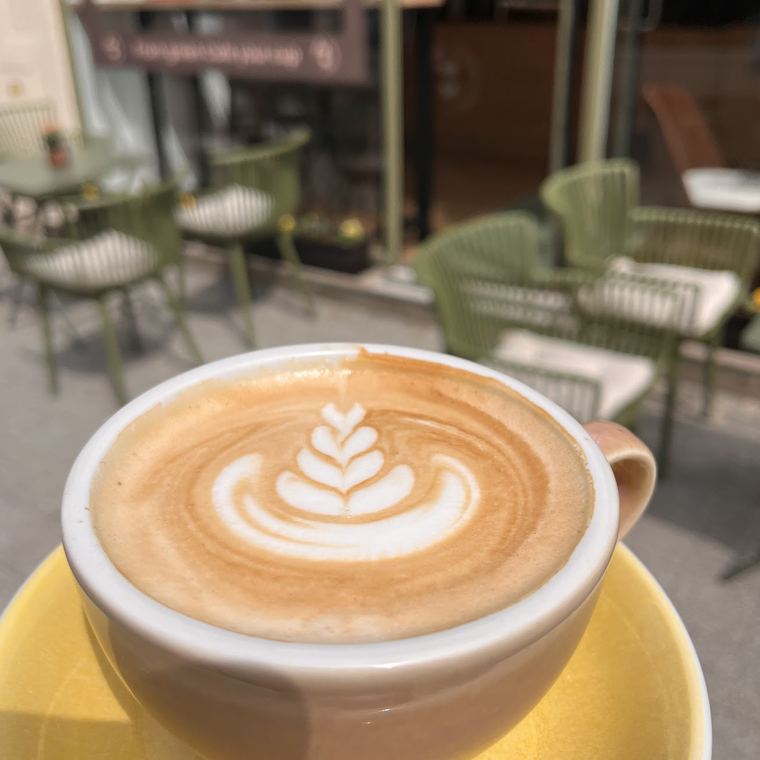 Cappuccino mit Latte Art in einer gelben Tasse, im Hintergrund die grünen Stühle des Gastgartens