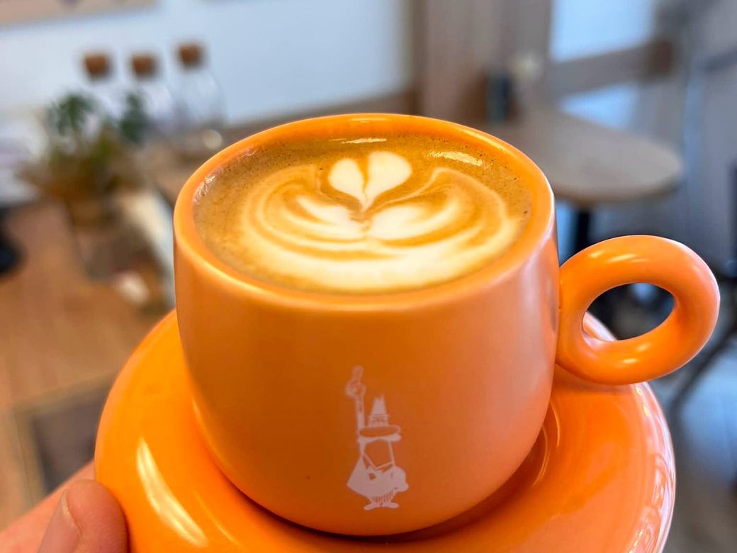 Cappuccino mit Latte Art in einer orangen Tasse des Hauses, dahinter der Innenraum mit den Kaffee-Illustrationen an der Wand