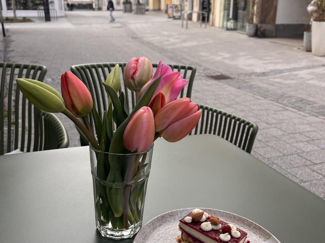 Ein Stück Schokoschnitte auf weißem Teller neben einem Tulpenstrauß auf dem salbeigrünen Tisch im Gastgarten